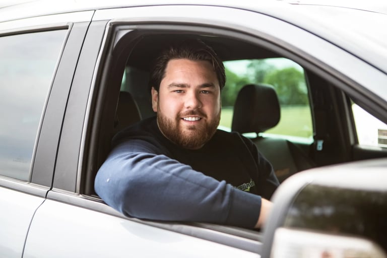 Bearded man smiling while sitting in drivers seat of white car, looking out open window