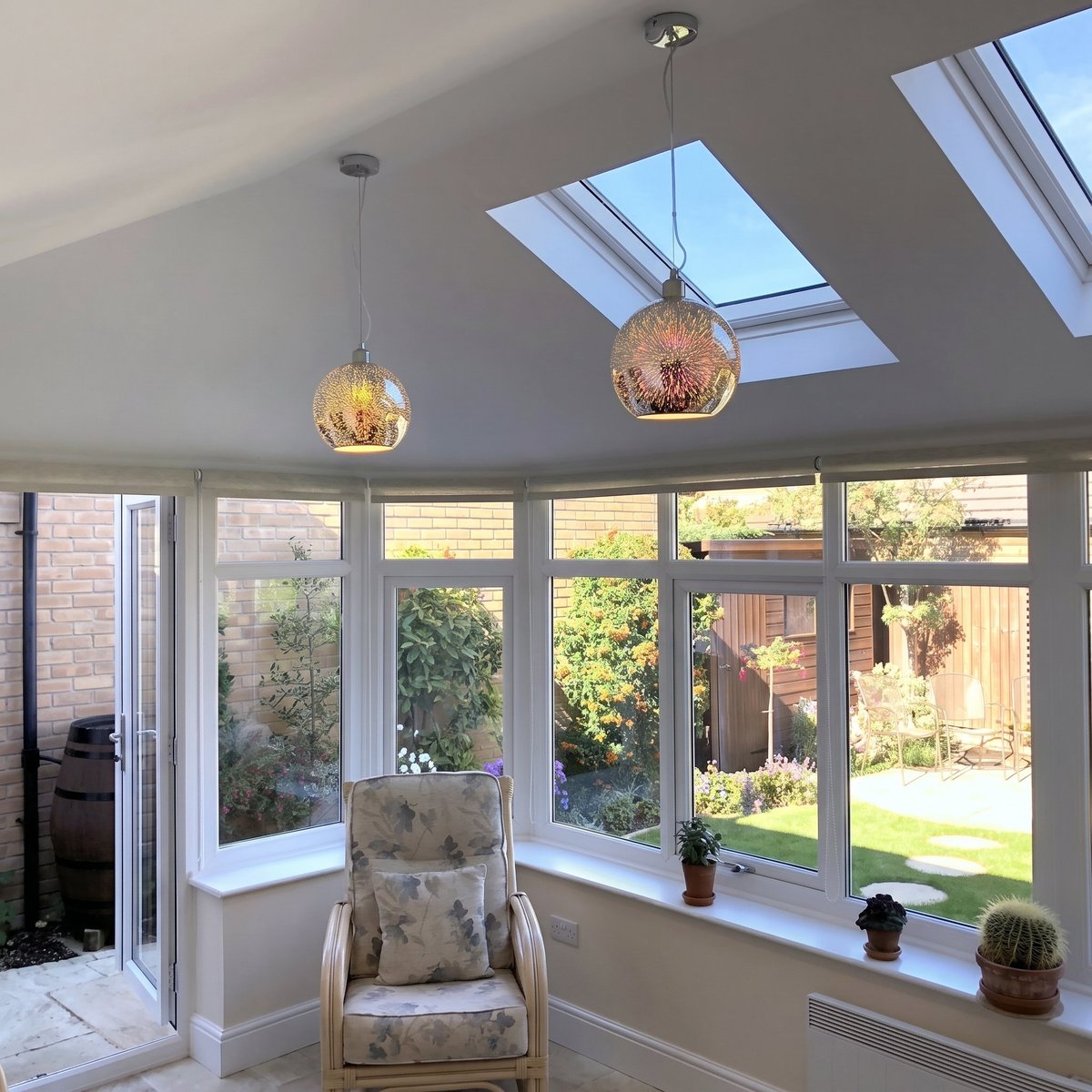 Modern sunroom with skylights, hanging pendant lights, large windows overlooking garden, and a cream armchair with potted plants on windowsill