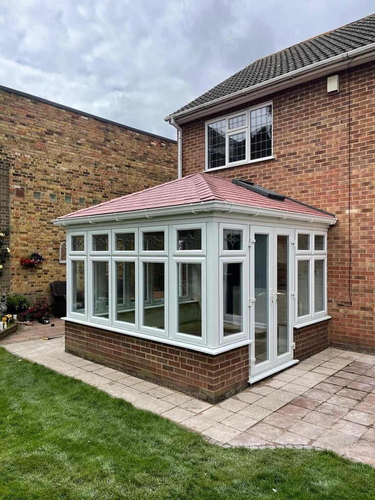 Modern white uPVC conservatory with pink roof attached to red brick house, featuring large windows and patio base.