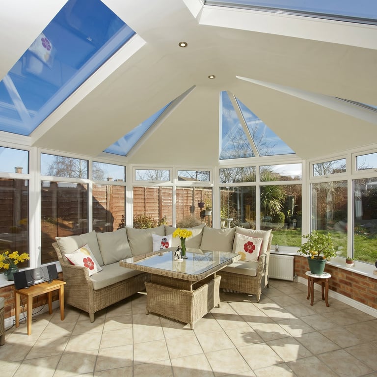 Bright sunlit conservatory with wicker furniture, cushions, skylights, and garden views through large windows