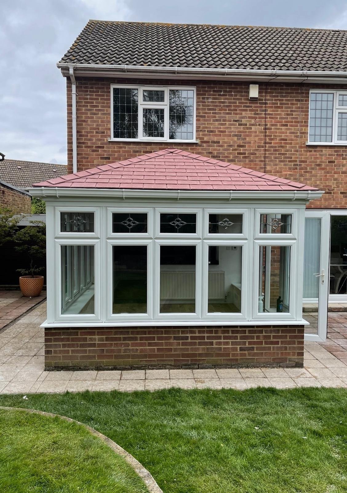 Modern brick house with white-framed glass conservatory featuring a pink pyramid roof and brick base, surrounded by patio and lawn