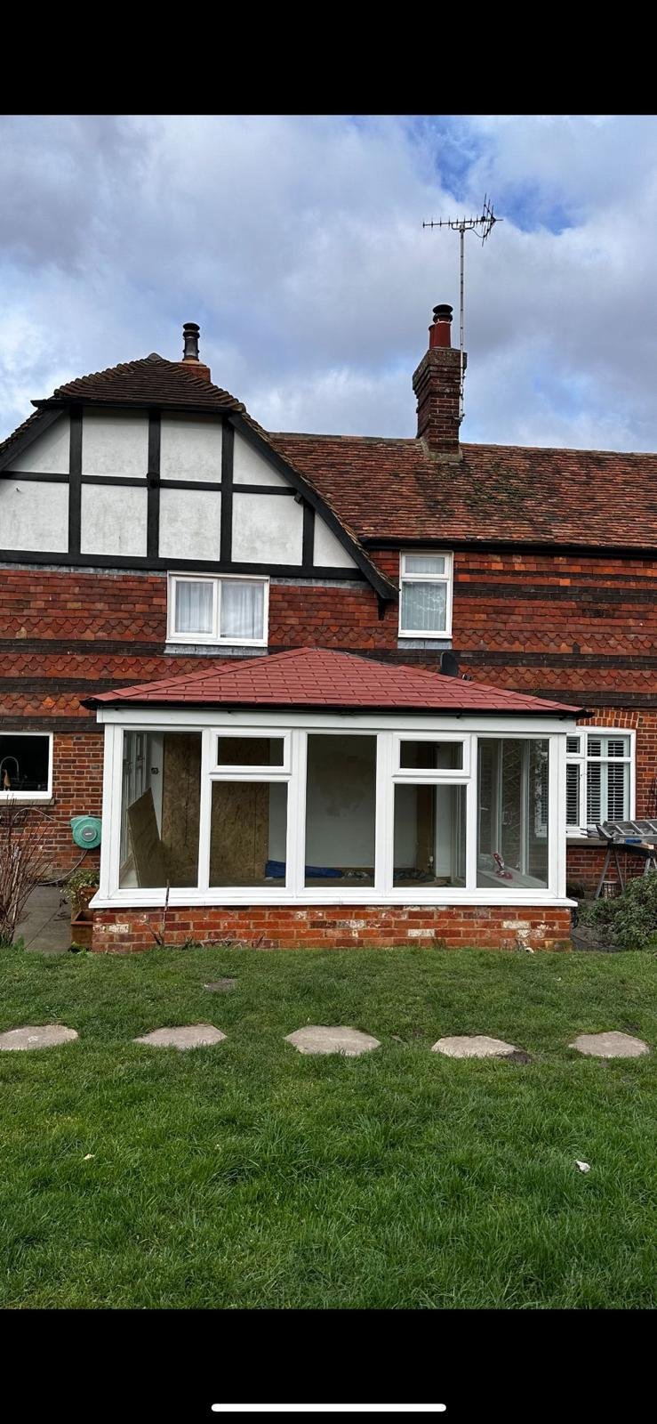 Tudor-style brick house with white-framed gable, modern conservatory, green lawn with stepping stones, and antenna on roof